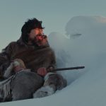 Benjamin Kunuk as Kuanana, seated low looking over a snowdrift into the distance with sunlight on his face, holding a rifle in his lap. Still from Maliglutit via The Cinematheque website, PHOTO COURTESY KINGULIIT PRODUCTIONS INC.