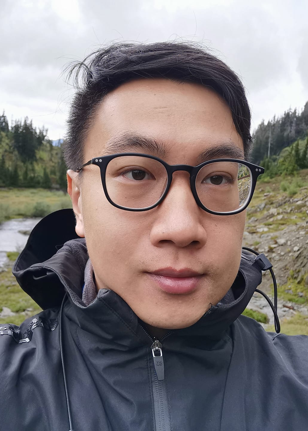 Portrait photo of Andrew Han, UBC alumnus (BA'14). Andrew is wearing glasses and a black waterproof jacket. His background indicates that he is outdoors.