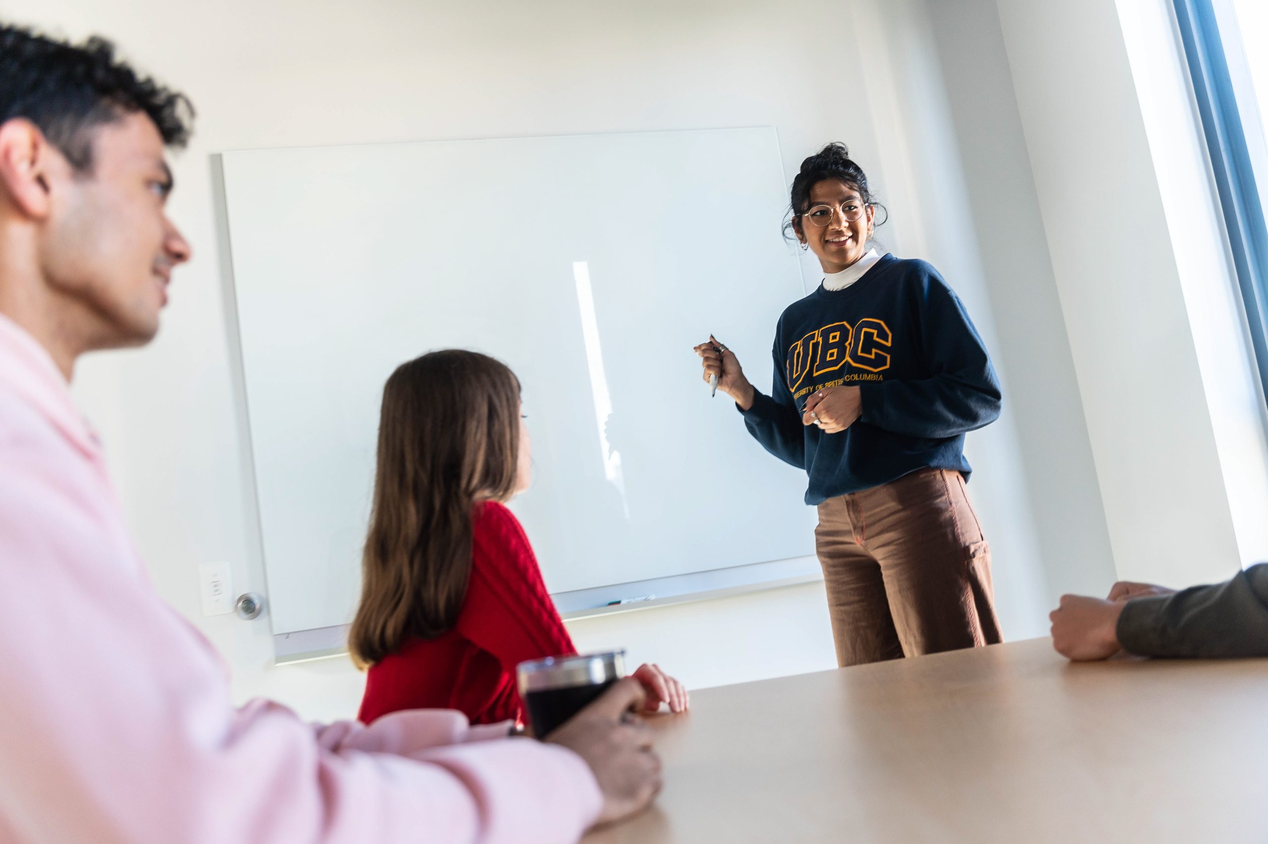 A student wearing a UBC sweatshirt stands at a whiteboard with a group in a study room.