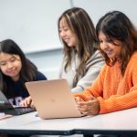 Three student in class at a table with their laptops.